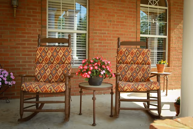 Two wooden rocking chairs with patterned cushions on a porch in front of a brick wall with two windows. A small round table with a pot of pink flowers is placed between the chairs. Additional potted plants are visible on the porch.