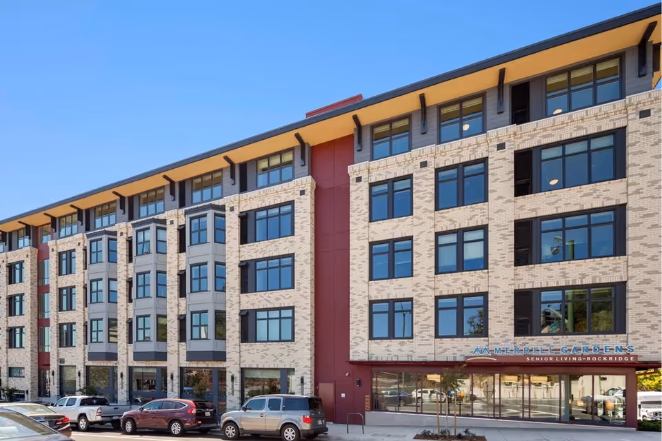 Exterior view of a modern multi-story senior living facility building with large windows and a parking area in front. The building has a beige brick facade with red and gray accents under a clear blue sky.