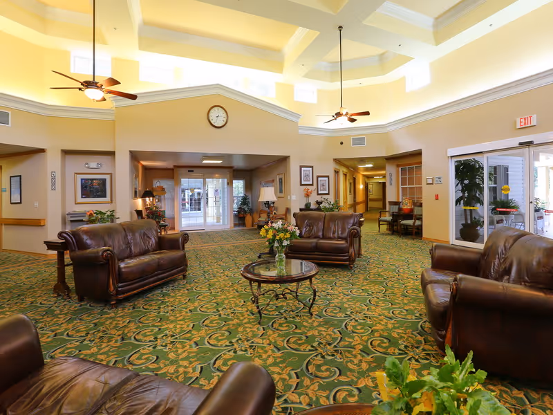 A spacious and well-lit common area with high ceilings and ceiling fans. The room features multiple brown leather sofas arranged around a glass coffee table with a flower vase. The floor is covered with a green patterned carpet. There are framed pictures on the walls and large windows and glass doors letting in natural light.