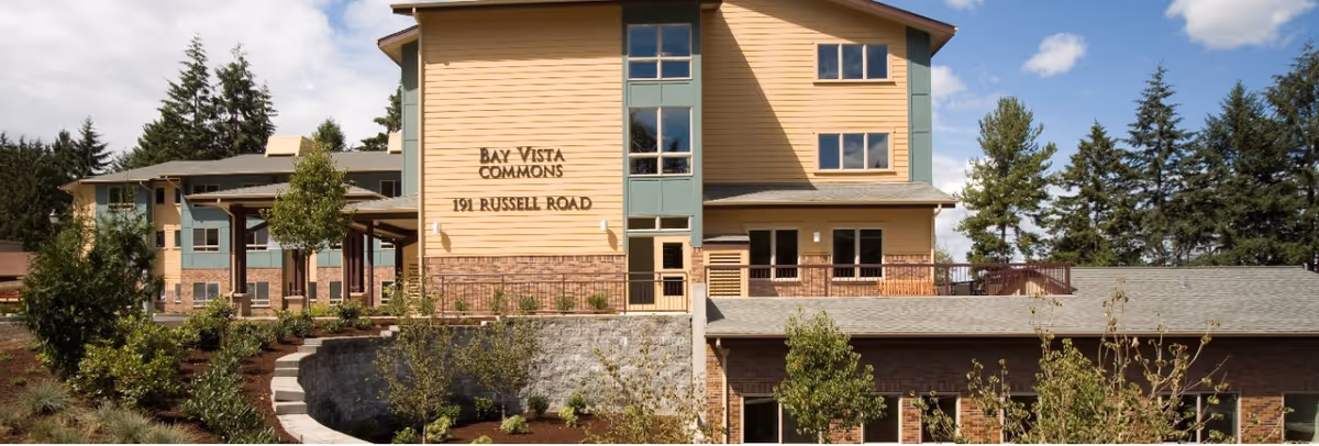 Exterior view of Bay Vista Commons building at 191 Russell Road, showing a multi-story structure with beige and green siding, surrounded by landscaped greenery and trees under a partly cloudy sky.