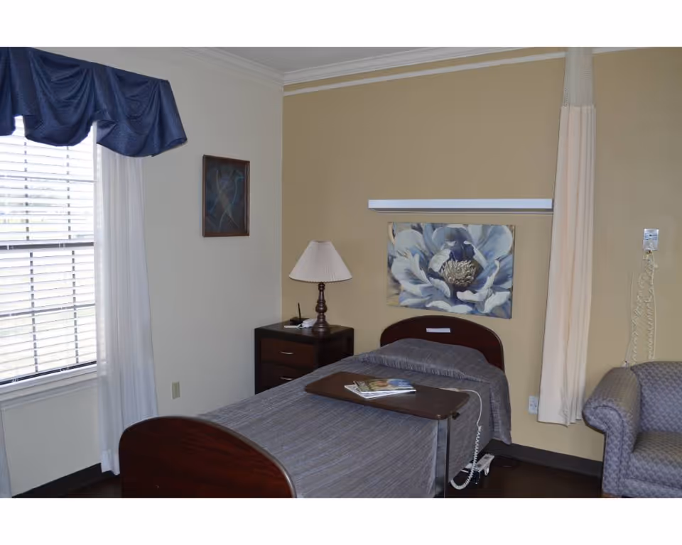 A single bed with a gray bedspread in a room with beige and white walls. There is a wooden nightstand with a lamp beside the bed, a floral painting above the headboard, a window with white curtains and a blue valance, and a gray upholstered armchair. A hospital-style overbed table with magazines is positioned over the bed, and a privacy curtain is partially visible on the right side.