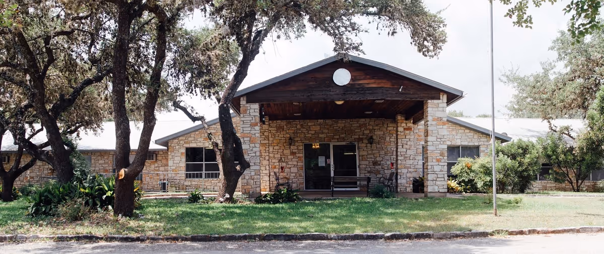 Stone-clad single-story building front with a covered entrance/porte-cochere, a flagpole, lawn and oak trees.