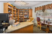 Interior view of a cozy library or reading room in a retirement community. The room features wooden bookshelves filled with books along the walls, a desk with a computer and office chair, and a small round table with two chairs near a window with blinds and valances. The ceiling has fluorescent lighting and the floor is carpeted.