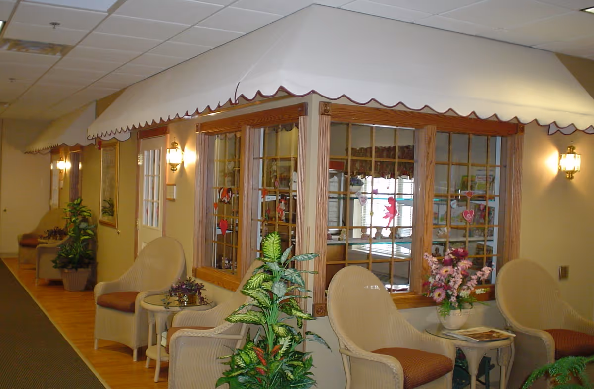 Indoor hallway seating area with wicker chairs and small tables adorned with plants and flowers. The walls have windows with wooden frames and white awnings above them. Warm lighting fixtures are mounted on the walls, creating a cozy atmosphere.