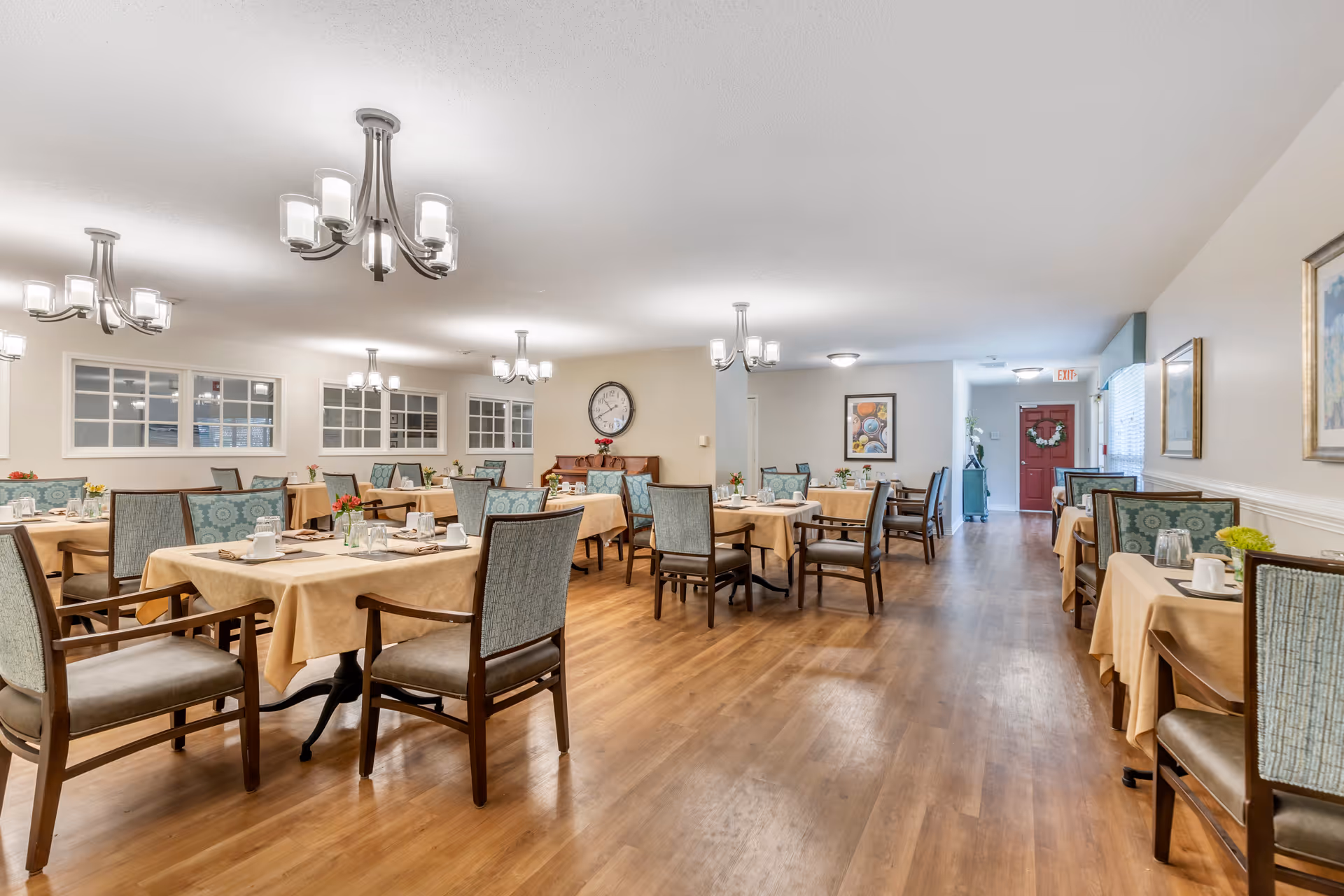 A spacious dining room with multiple tables covered in beige tablecloths, each set with cups, plates, and small flower arrangements. The room has wooden flooring, several chandeliers hanging from the ceiling, and framed artwork on the walls. There is a red door at the far end of the room and large windows on one side.