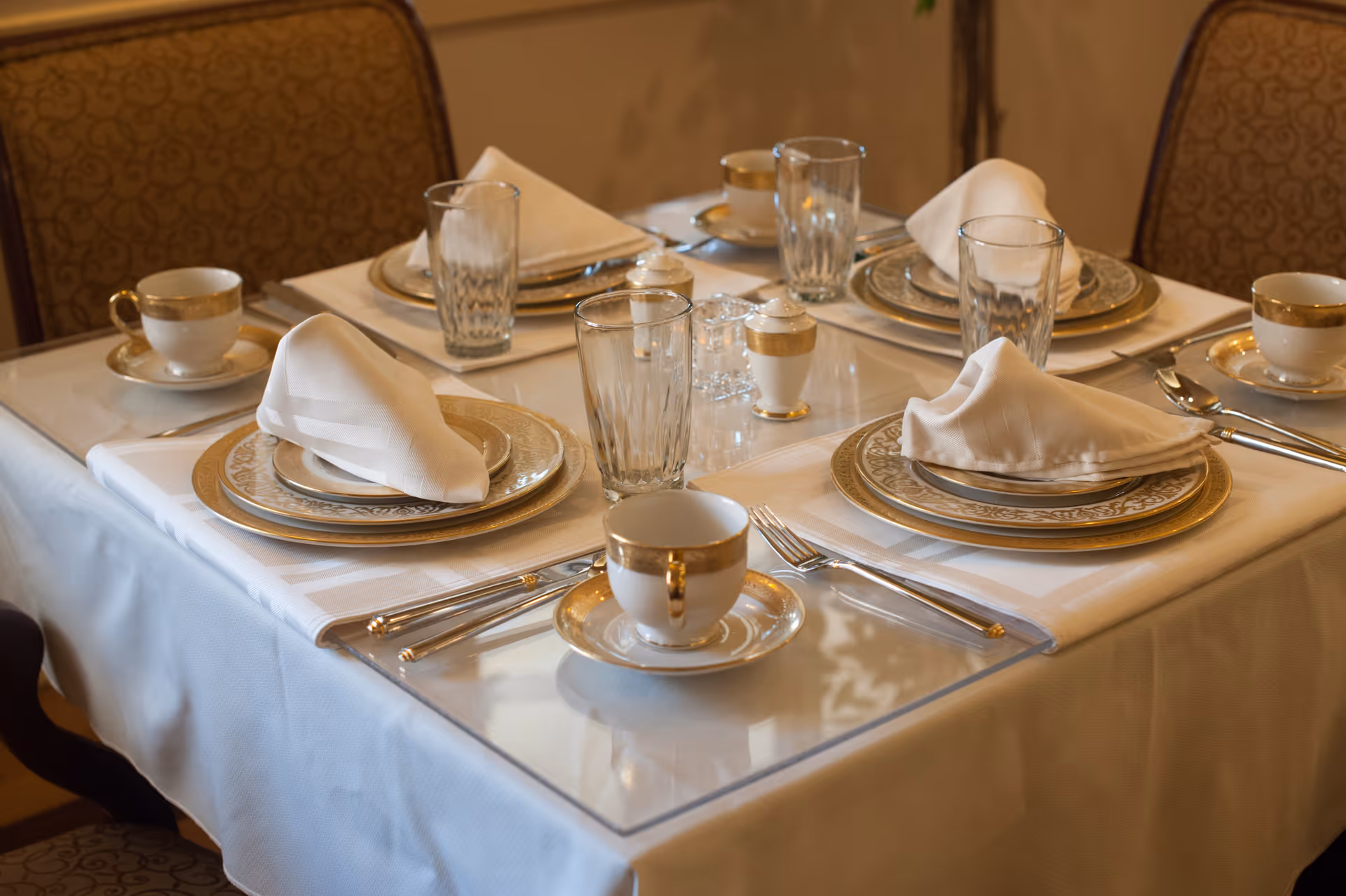 A dining table set for four with white tablecloth, gold-rimmed plates, cups, and saucers, folded white napkins, clear drinking glasses, and silverware arranged neatly on placemats.