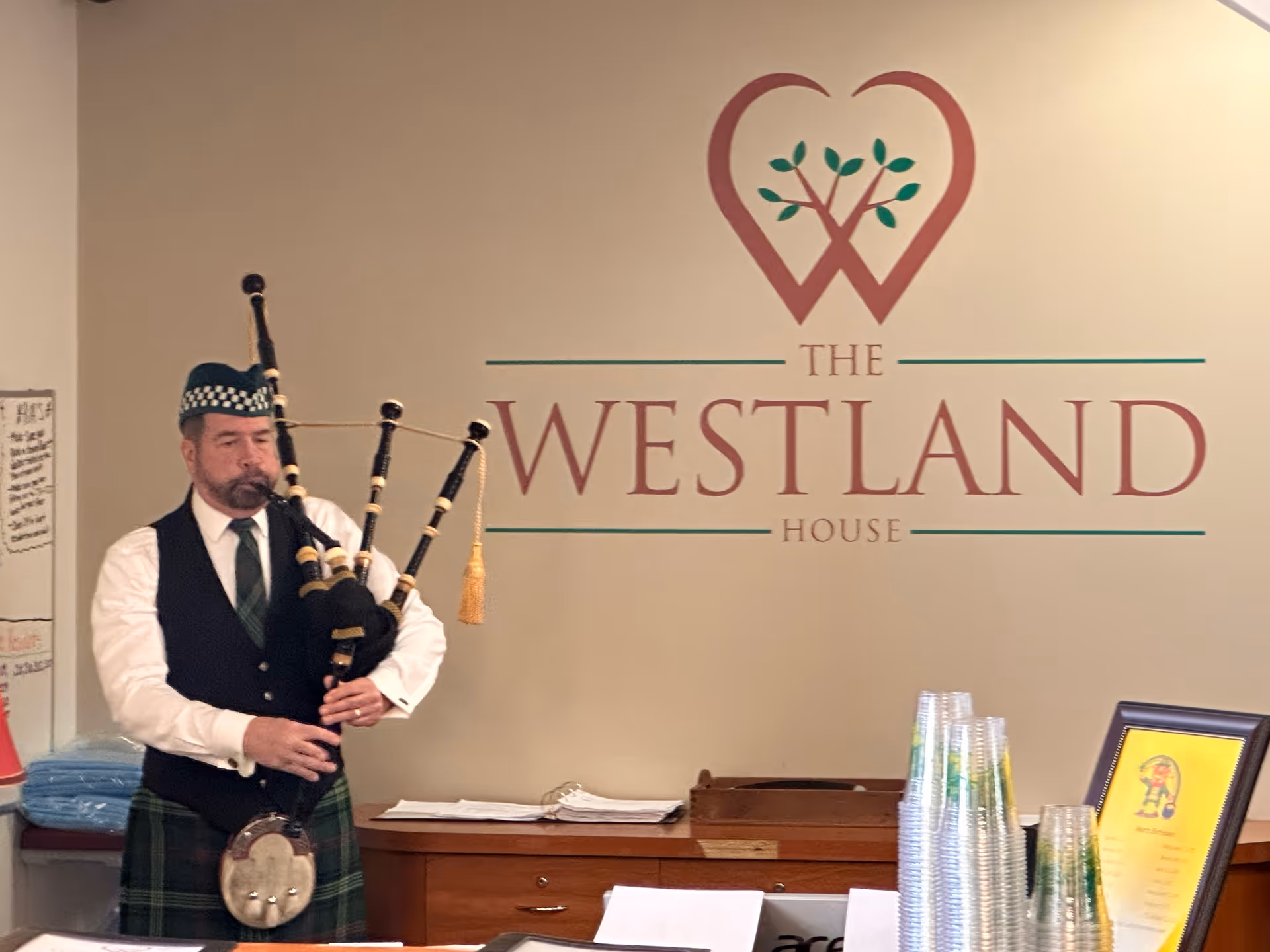 A man dressed in traditional Scottish attire playing bagpipes indoors in front of a wall with the logo and name 'The Westland House'. There are stacked plastic cups and a framed certificate on a counter nearby.