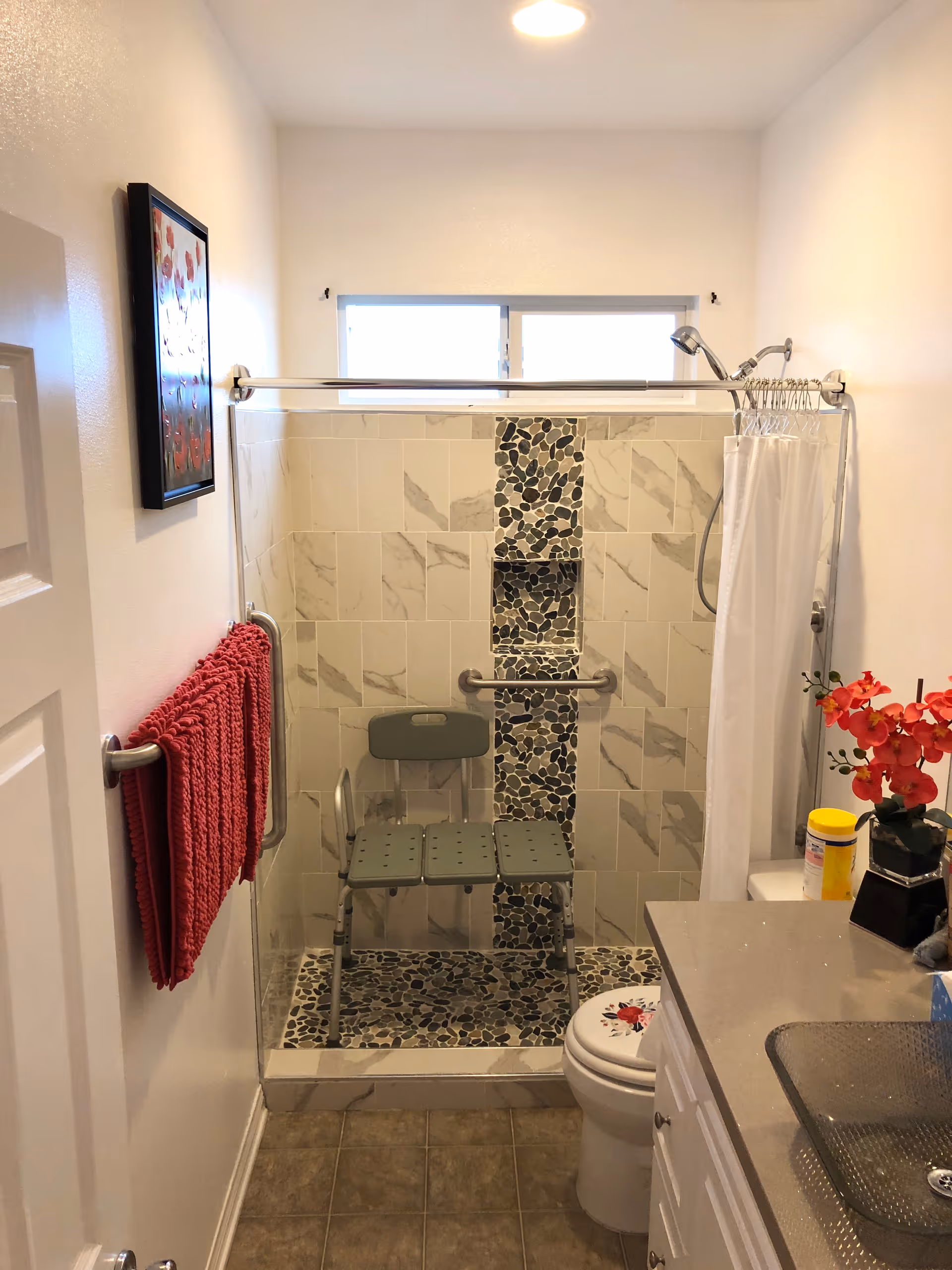 A small bathroom with a walk-in shower featuring a pebble stone and tile design. Inside the shower is a green shower chair and a grab bar. A red towel hangs on a towel rack on the left wall. There is a white toilet with a floral seat cover next to the shower. On the right, a countertop with a glass sink, a container of disinfecting wipes, and a vase with red flowers is visible. A framed picture hangs on the left wall, and a window above the shower provides natural light.