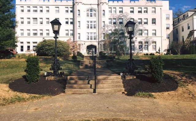 Front exterior view of a large, multi-story white building with numerous windows, a central entrance with steps leading up, flanked by two black vintage-style street lamps and landscaped bushes on either side.