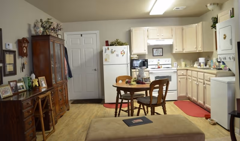 Interior view of a senior living apartment kitchen and dining area featuring light wood cabinets, white appliances including a refrigerator, stove, microwave, and stacked washer and dryer. A small round wooden dining table with two chairs is centered in the room. To the left, there is a wooden cabinet with framed photos and decorative items. The floor is light wood, and the walls are painted beige.