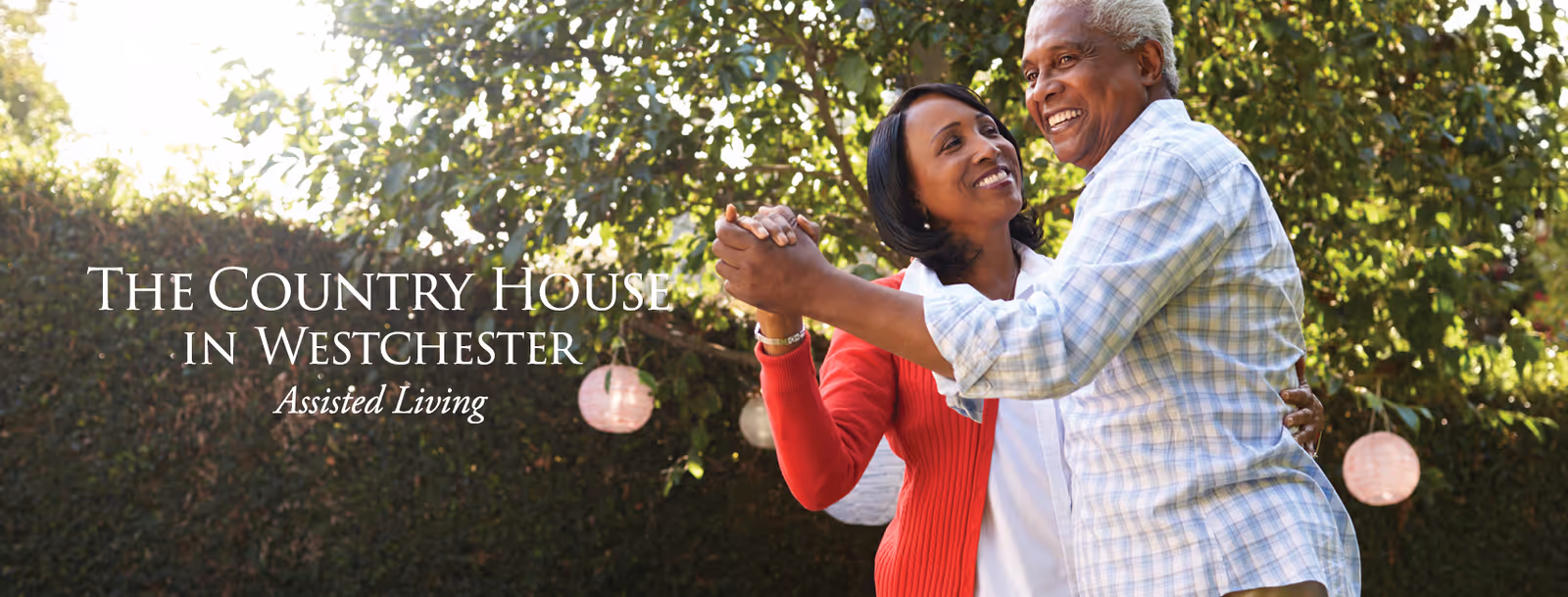 An elderly couple dancing happily outdoors in a garden with trees and hanging lanterns in the background. The man is wearing a light blue checkered shirt and the woman is wearing a red cardigan over a white top. Text on the image reads 'The Country House in Westchester Assisted Living'.