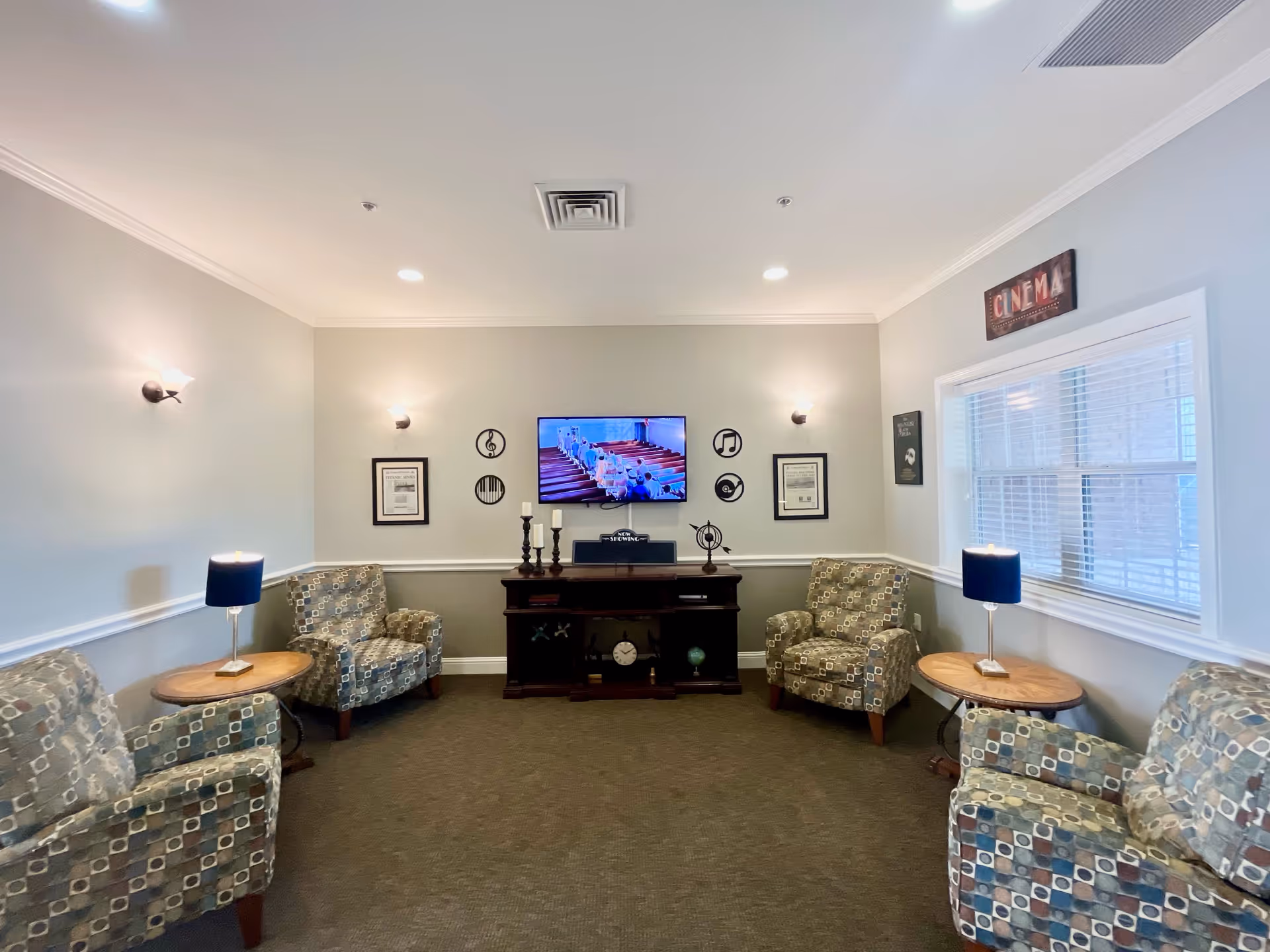 A cozy living room with four patterned armchairs arranged around two wooden side tables with blue lamps. A flat-screen TV is mounted on the wall above a dark wooden console table decorated with candles and small ornaments. The walls are light gray with white trim, and there are musical-themed decorations and framed pictures on the wall. A window with white blinds is on the right side, and a sign above it reads 'CINEMA'.