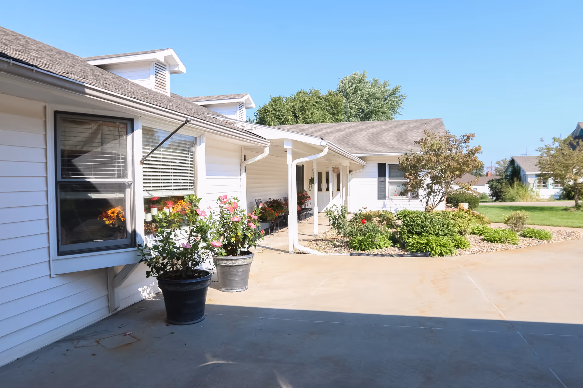 Front exterior of a single-story white building with potted flowers, a covered entryway, and landscaped beds under a clear blue sky.