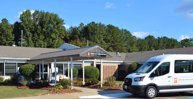 Exterior view of Christian Care Center showing a single-story building with a covered entrance, well-maintained landscaping including bushes and flowers, and a white van parked in front with the facility's name on it. Trees and a blue sky with some clouds are visible in the background.