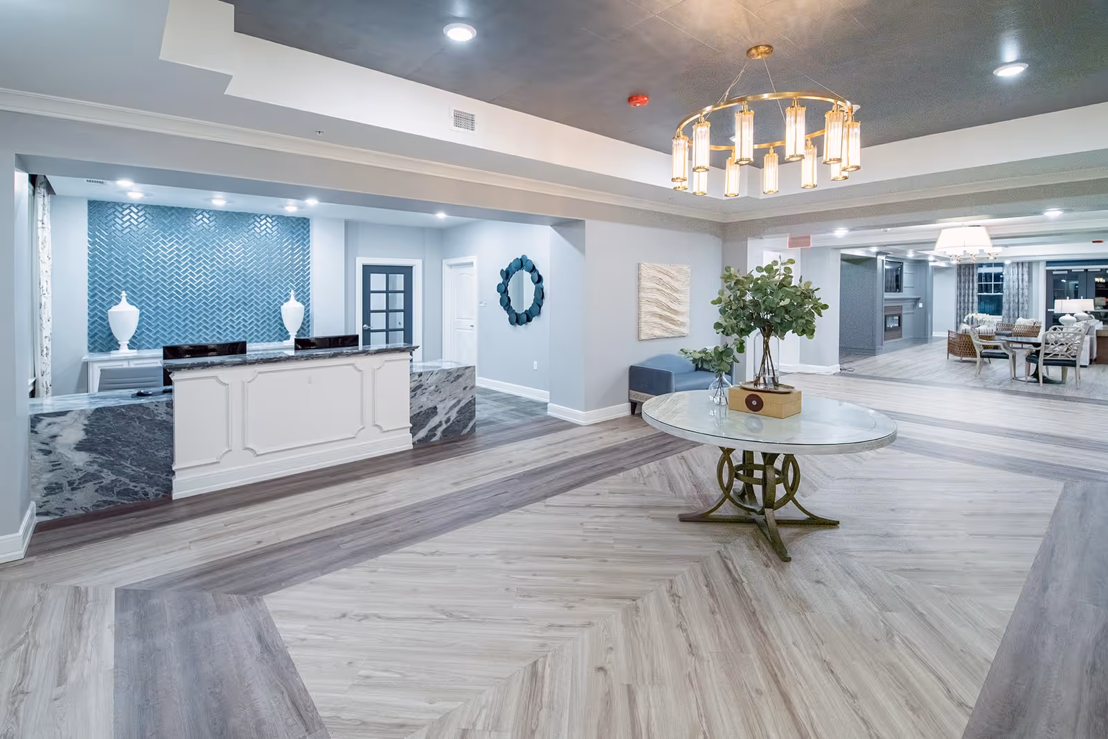 A spacious and modern senior living facility lobby with a reception desk featuring a marble countertop and a blue textured wall behind it. The floor has a stylish wood pattern, and a round table with a vase of greenery is centered under a decorative chandelier. In the background, there is a seating area with chairs and a fireplace.