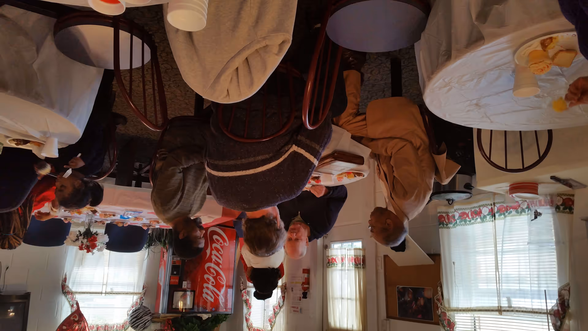 A group of elderly people sitting around tables in a dining area, engaging in conversation and eating. The room has windows with floral curtains, a Coca-Cola vending machine, and several chairs and tables covered with white tablecloths.