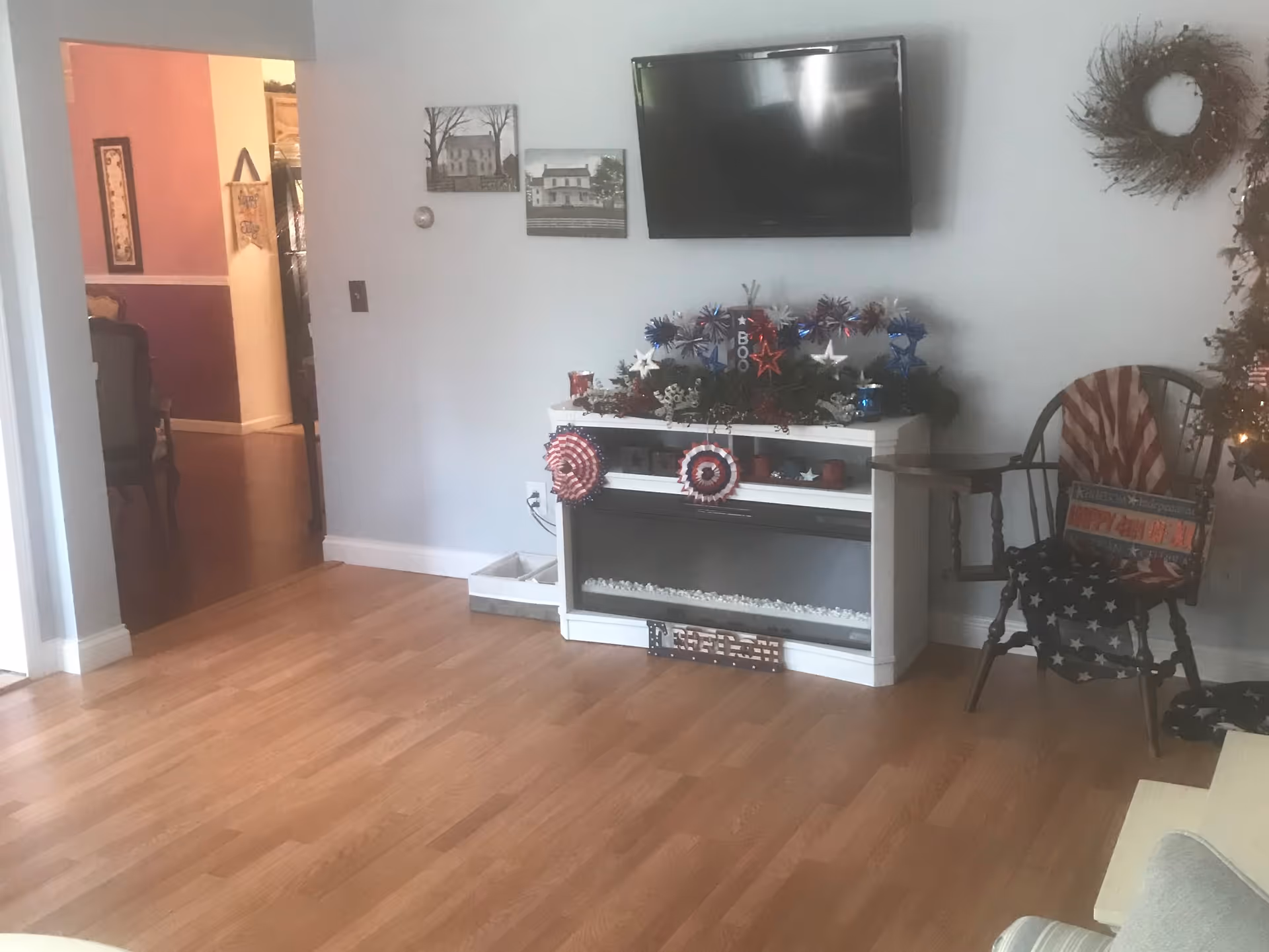 A living room with light wood flooring, a white electric fireplace decorated with patriotic red, white, and blue ornaments, and a flat-screen TV mounted on the light gray wall above it. To the right, there is a wooden chair draped with an American flag-themed blanket and a decorative sign. On the left, there is an open doorway leading to another room with purple and pink walls and a wooden dining chair partially visible.