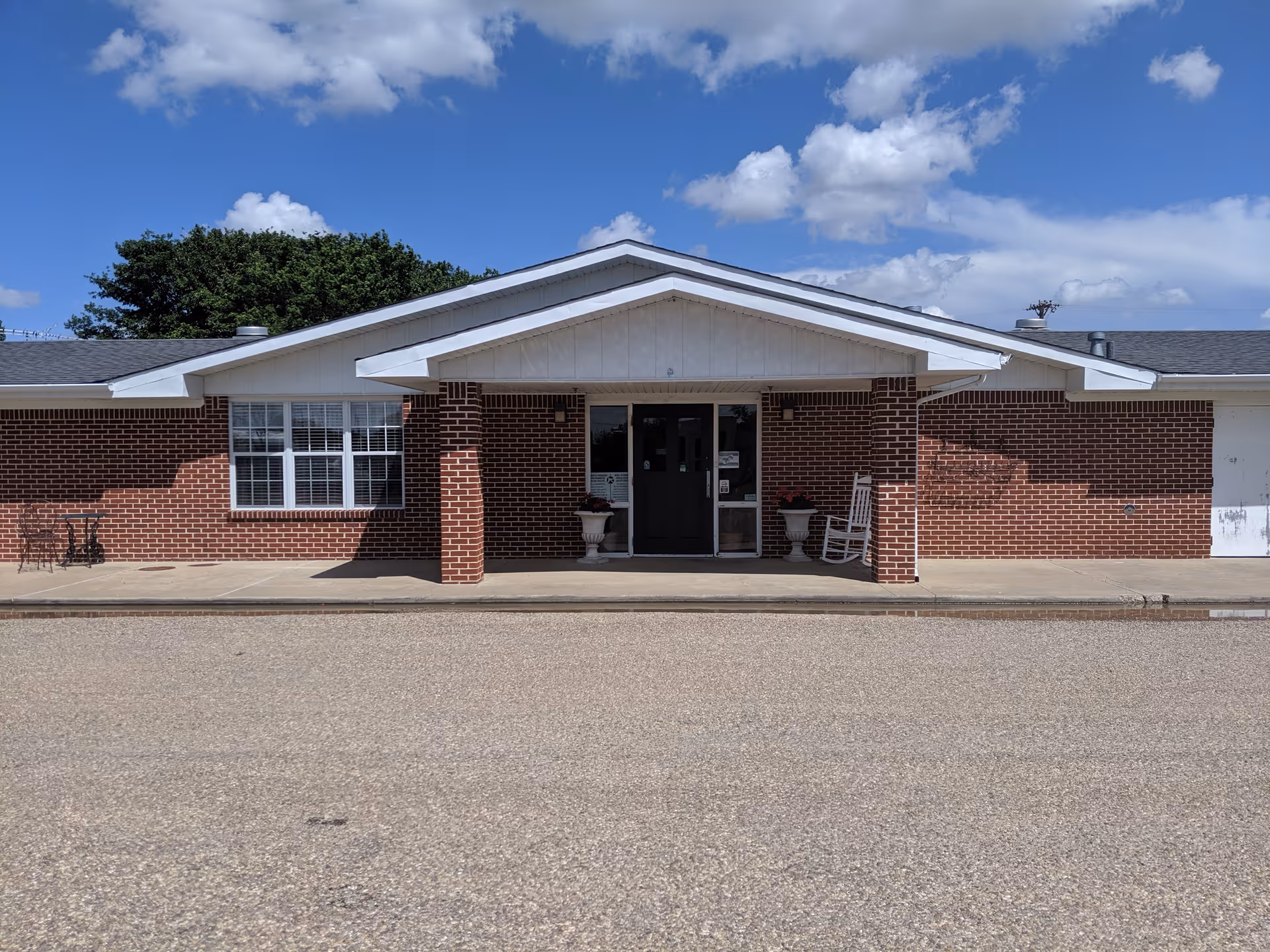 Front exterior view of a single-story brick building with a covered entrance supported by two brick columns. There are two white planters and a white rocking chair near the entrance door. The sky is blue with scattered clouds.