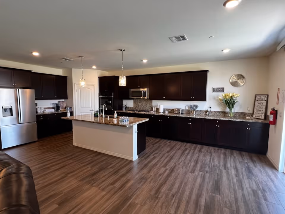 A spacious kitchen with dark wood cabinets and granite countertops. The kitchen features a large island with a sink and two pendant lights hanging above it. Stainless steel appliances including a refrigerator, microwave, and oven are visible. The floor is covered with wood-like vinyl planks. On the right side, there is a vase with yellow flowers, a wall clock, and a framed sign with kitchen rules. A fire extinguisher is mounted on the wall near the entrance.