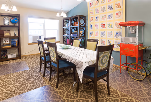 A dining room with a rectangular table covered with a white patterned tablecloth surrounded by six wooden chairs with decorative cushions. The room has patterned carpet flooring, a window with an air conditioning unit, two bookshelves filled with books and decorative items, a colorful quilt hanging on the wall, and a vintage-style red popcorn machine on wheels.