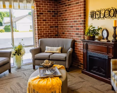 A cozy living room area with a gray loveseat, a matching armchair, and a round ottoman covered with a yellow throw blanket. There is a decorative tray with items on the ottoman. The room features a brick wall corner, a wooden electric fireplace with decorative items on top, and a 'WELCOME' sign on the wall. A large window with a yellow and white valance lets in natural light, and a vase with flowers is placed near the window.