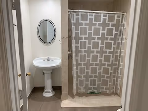 Small bathroom with a pedestal sink and oval mirror next to a shower with a patterned curtain.