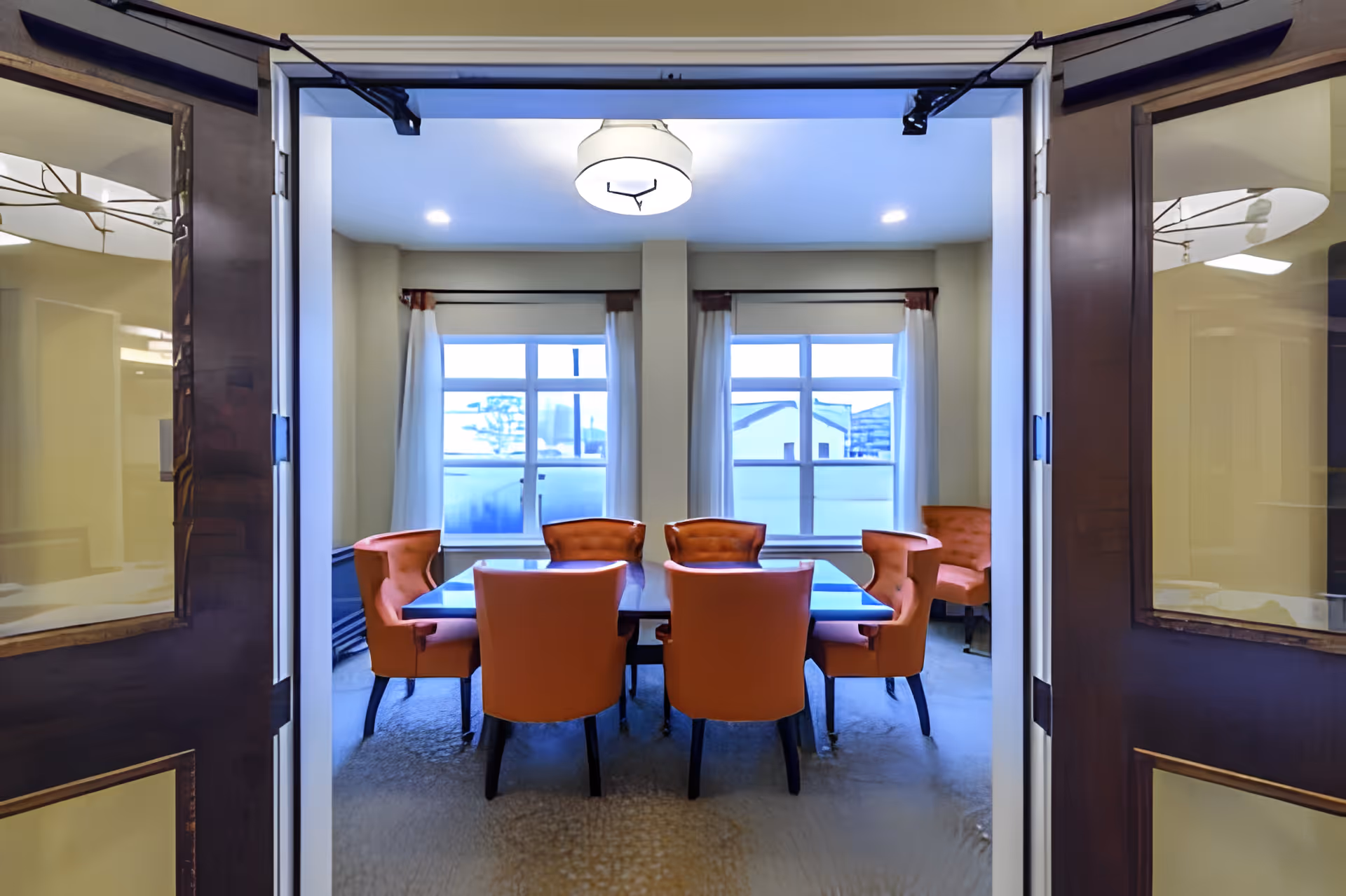 A dining room with a rectangular table surrounded by eight orange upholstered chairs. The room has two large windows with white curtains and a ceiling light fixture. The photo is taken from an open doorway with dark wooden doors.