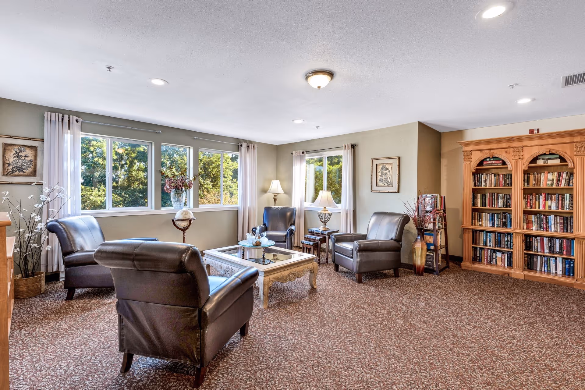 A cozy living room with four dark leather armchairs arranged around a glass-top coffee table. The room has large windows with white curtains letting in natural light, a wooden bookshelf filled with books, two table lamps on side tables, and decorative plants and framed artwork on the walls.