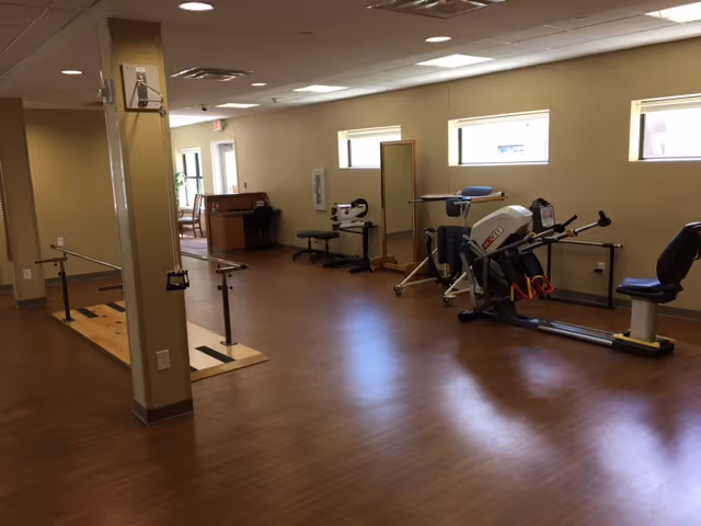 Well-lit interior therapy/fitness room with exercise equipment, parallel bars, and wood flooring.