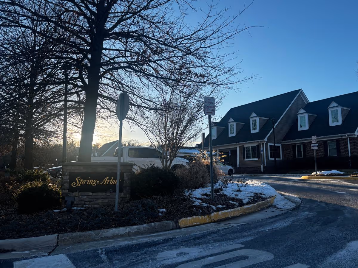 Entrance drive with a 'Spring Arbor' sign, parked cars, and a two-story brick building with snow on the ground under a clear sky.
