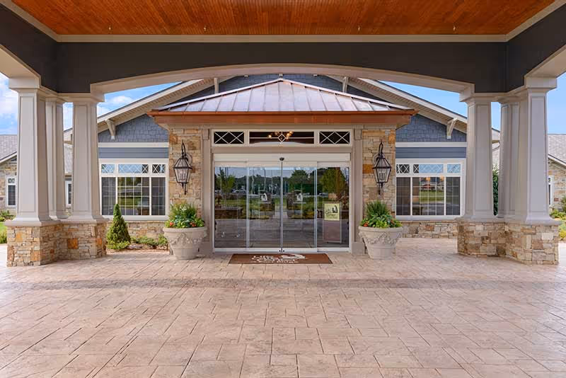 Covered porte-cochere leading to the glass double-door main entrance of a senior living building with stone columns and planters.
