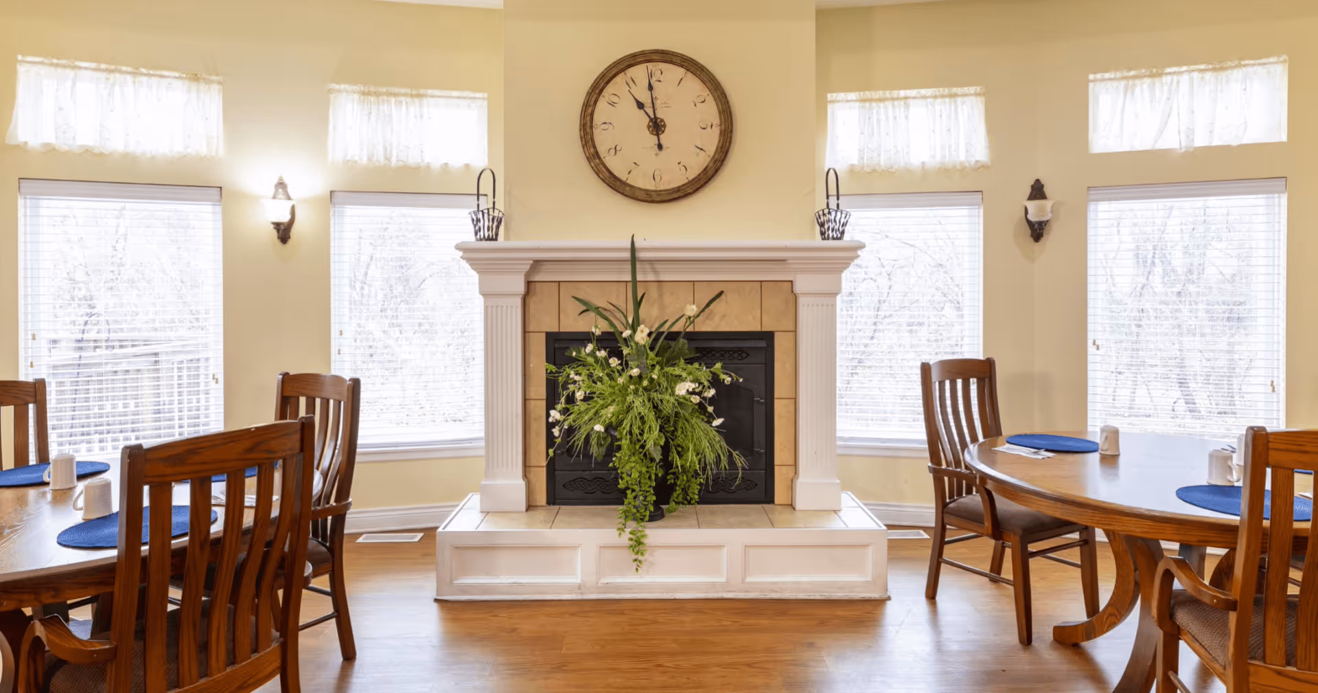 A bright dining room with wooden tables and chairs arranged around a central fireplace. The fireplace has a large round clock above it and a green floral arrangement on the hearth. Multiple windows with white blinds and lace valances allow natural light to fill the room.