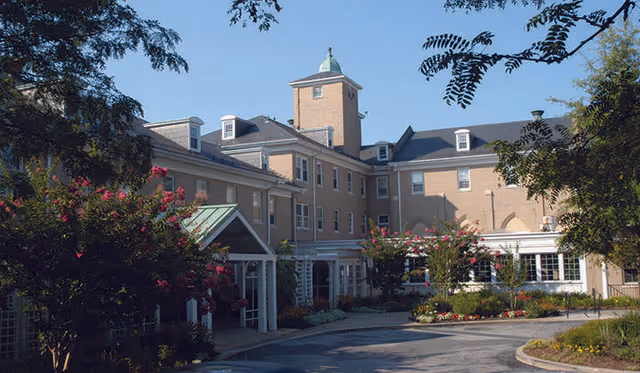 Exterior view of Glen Meadows Retirement Community building with a multi-story beige brick facade, a central tower with a green dome, surrounded by trees and flowering bushes under a clear blue sky.