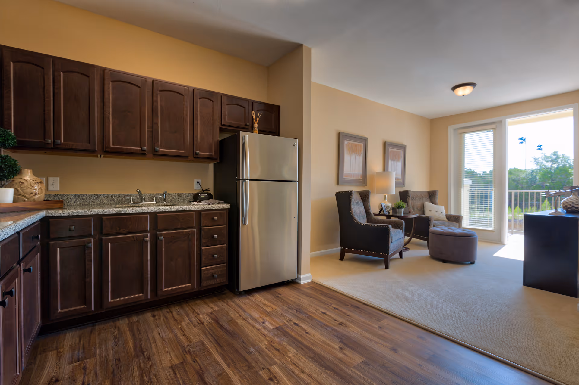 Interior view of a senior living facility showing a kitchen area with dark wooden cabinets, granite countertops, and a stainless steel refrigerator. Adjacent to the kitchen is a living room area with two upholstered armchairs, a small side table with a lamp, framed artwork on the wall, and a glass door leading to an outdoor balcony with greenery visible outside.