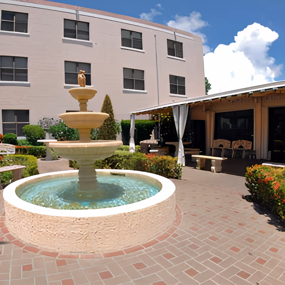 Outdoor courtyard area of a senior living facility featuring a three-tiered stone water fountain surrounded by greenery and bushes. There is a covered patio area with benches and chairs to the right, and a multi-story building with several windows in the background under a blue sky with some clouds.