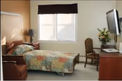 Small, bright resident bedroom with a single bed covered by a patchwork quilt, a window with valance, a wall-mounted TV, chair, lamp, and dresser with flowers.