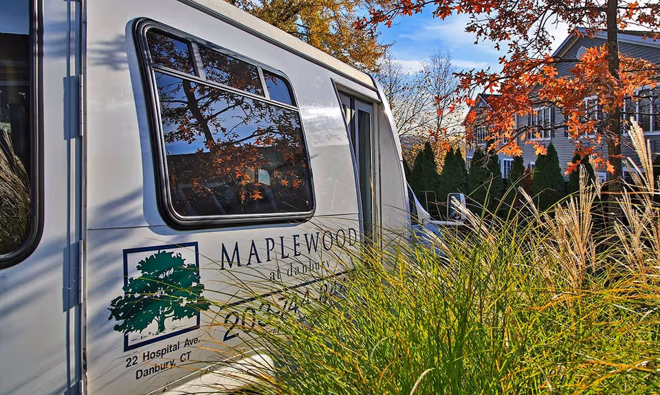 Side view of a white vehicle with the logo and name 'Maplewood at Danbury' parked near tall grass and autumn trees with orange leaves. A residential building is visible in the background under a clear blue sky.