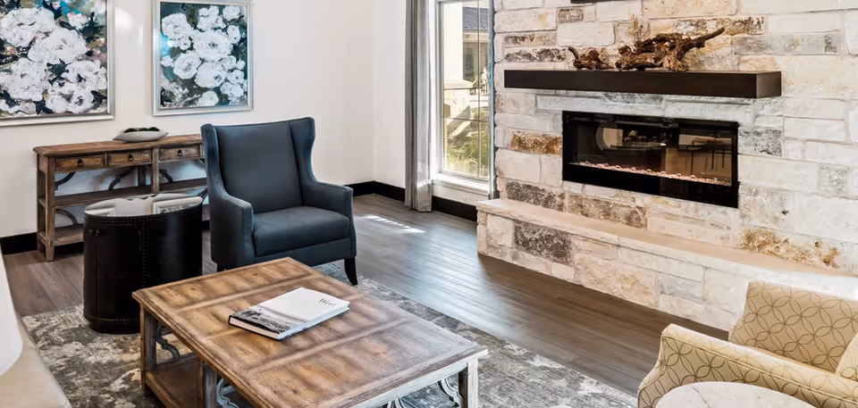 A cozy living room area featuring a stone fireplace with a modern electric insert, a blue upholstered armchair, a wooden coffee table with a book on it, a round black side table, a wooden console table against the wall, two floral paintings, and a large window with curtains letting in natural light.