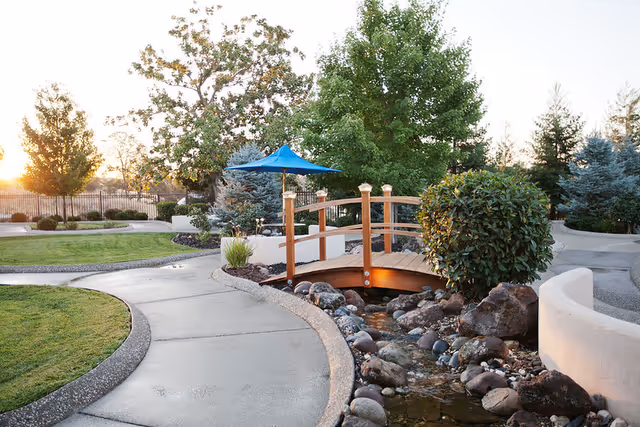 A serene outdoor garden area at Westview Healthcare Center featuring a small wooden footbridge over a rocky stream, surrounded by green trees, bushes, and a paved walking path. A blue umbrella is visible in the background providing shade.