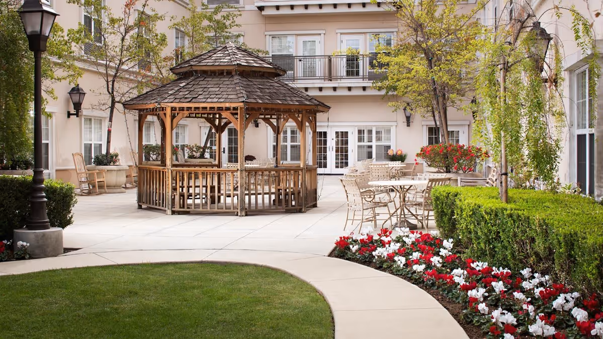 Outdoor courtyard area at Belmont Village Senior Living Encino featuring a wooden gazebo with seating inside, surrounded by patio tables and chairs. The courtyard is landscaped with green bushes, red and white flowers, and small trees, with a building facade in the background.