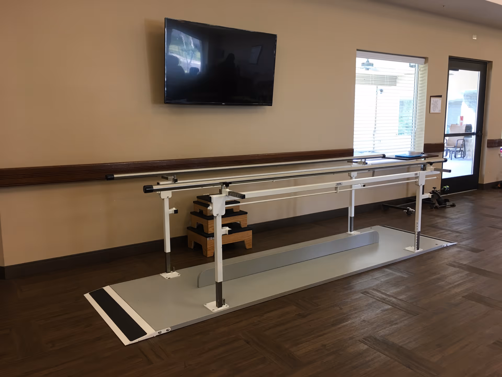 Parallel bars and a step platform in a rehabilitation room with a wall-mounted TV and a windowed door.