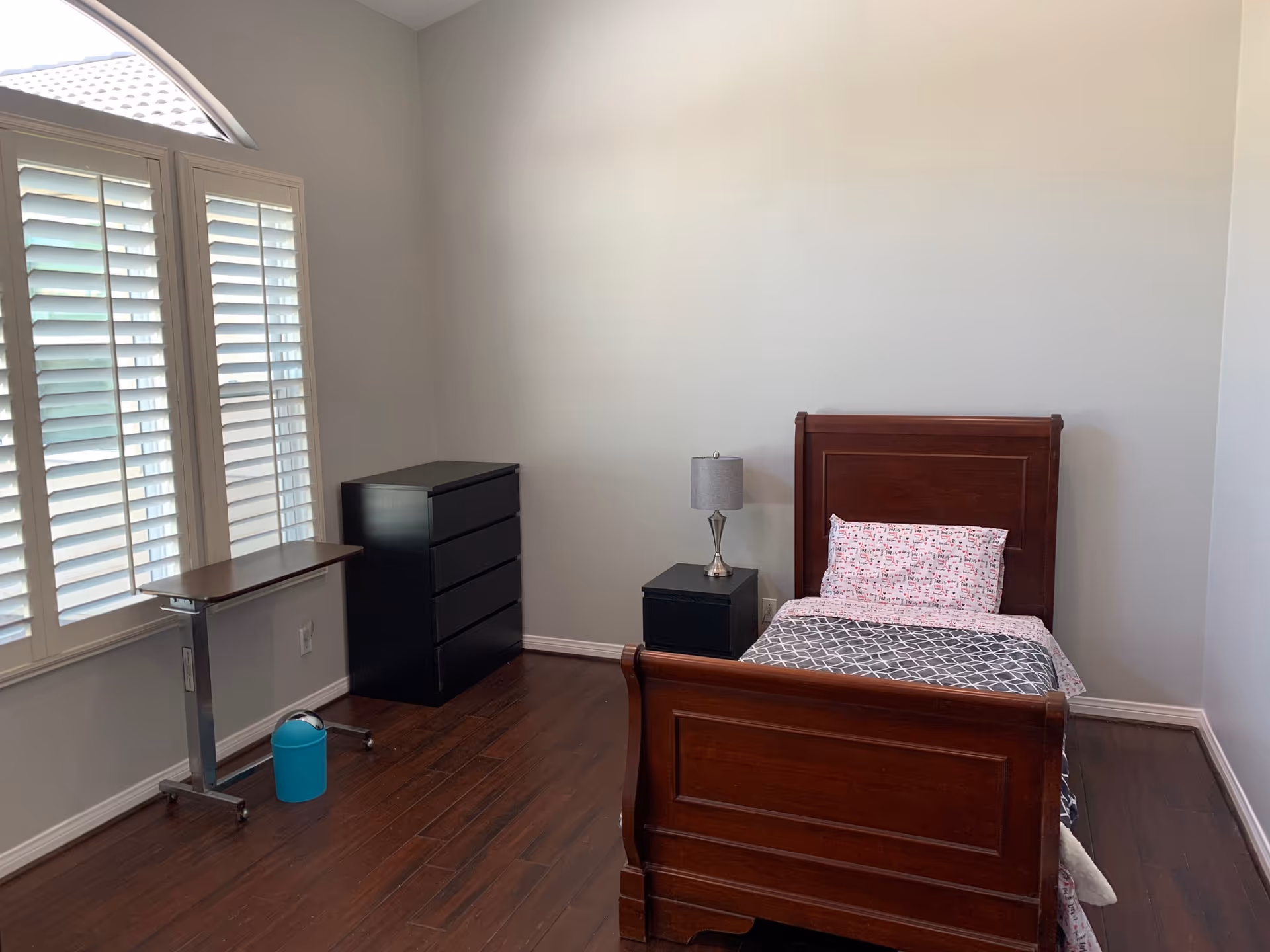 Minimal bedroom with a single wooden bed, nightstand and lamp, black dresser, overbed table, and shuttered windows.