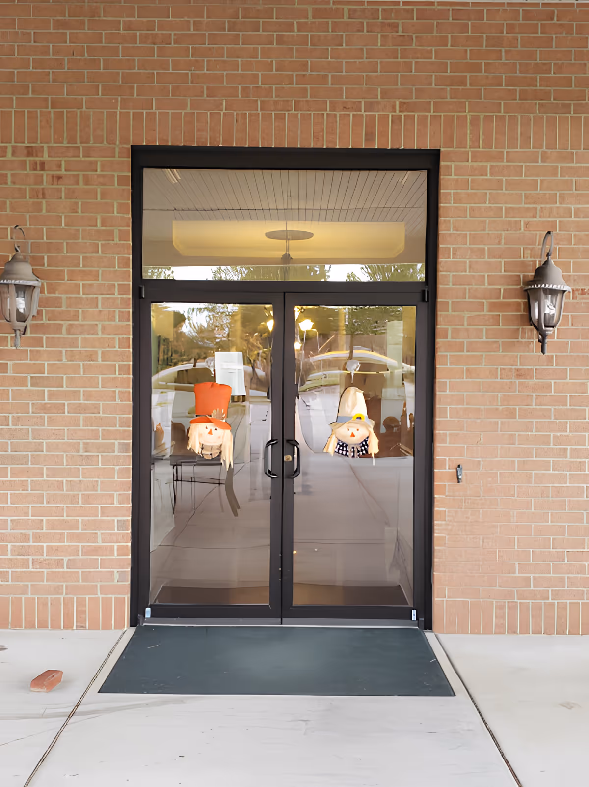 Double glass doors with black frames set in a brick wall. Each door has a scarecrow decoration hanging on it. There are two wall-mounted lantern-style lights on either side of the doors. The ground in front of the doors is concrete with a dark mat.