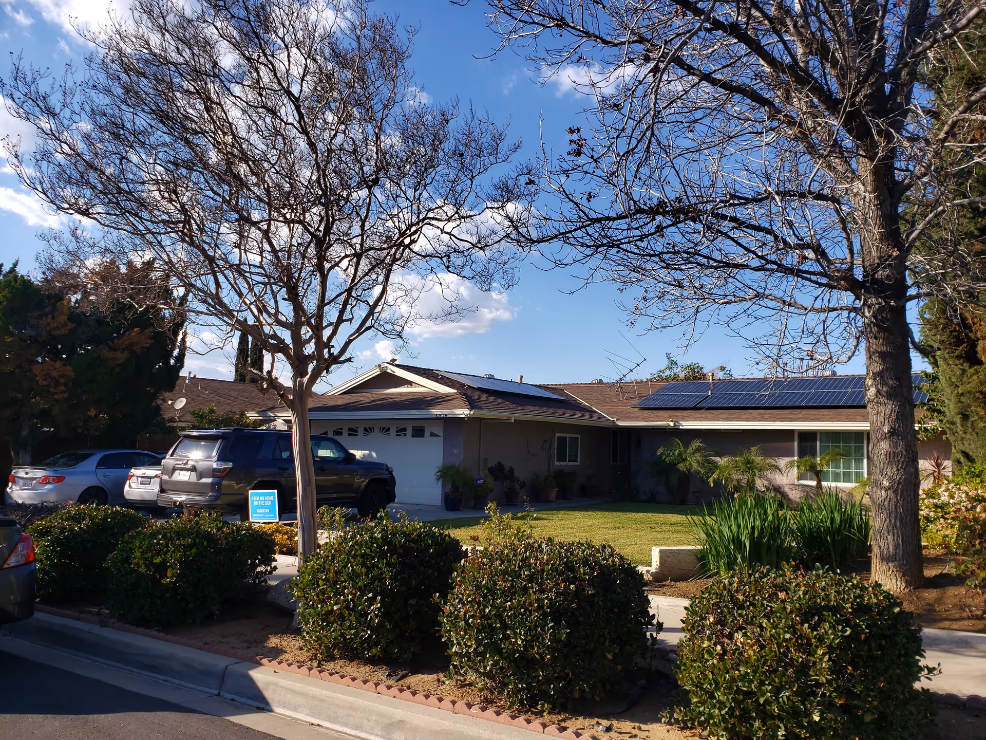 Front exterior view of a single-story residential building with a garage, solar panels on the roof, several parked cars, leafless trees, and well-maintained bushes and lawn under a partly cloudy blue sky.