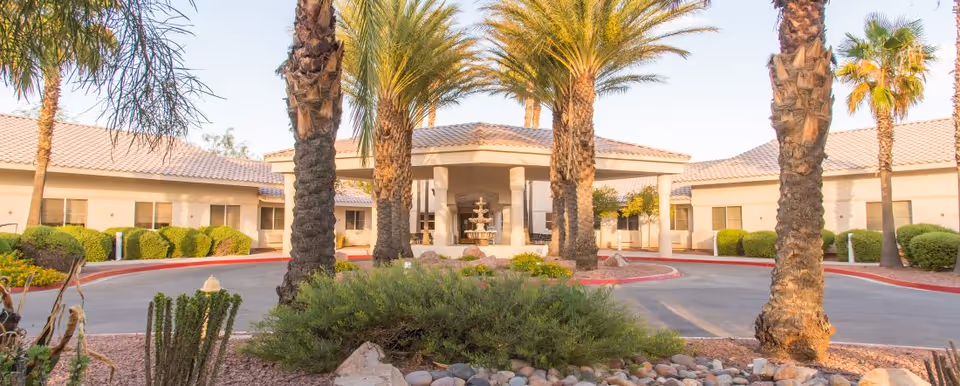 Front entrance of a single-story senior living building with a covered portico, central fountain, circular driveway, and palm trees.