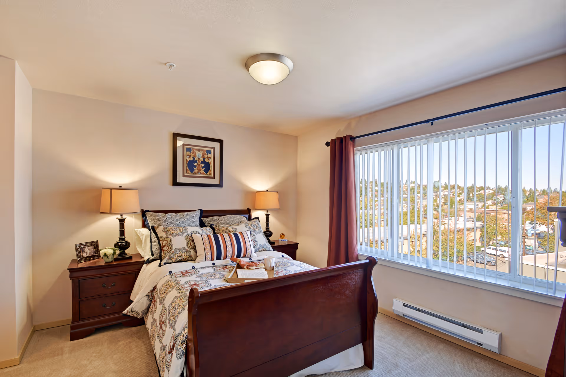 A cozy bedroom with a wooden bed frame and a bed made with patterned bedding and multiple pillows. There are two bedside tables with lamps and a framed picture on the wall above the bed. A large window with vertical blinds and maroon curtains lets in natural light and offers a view of a neighborhood outside.