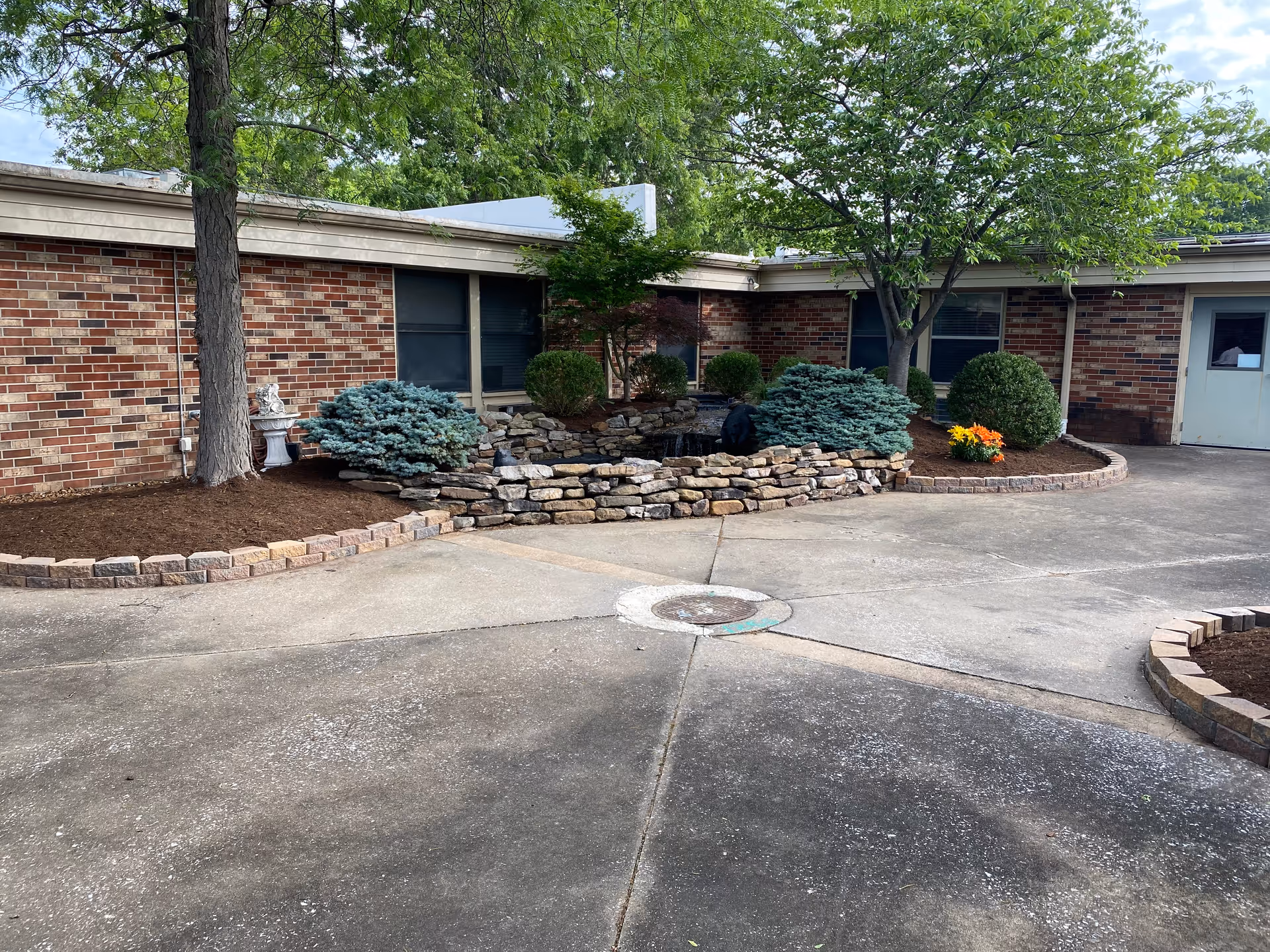 Outdoor courtyard area of a brick building with a small landscaped garden featuring shrubs, a tree, a stone retaining wall, and a small water feature. The ground is paved with concrete, and there is a door visible on the right side of the building.