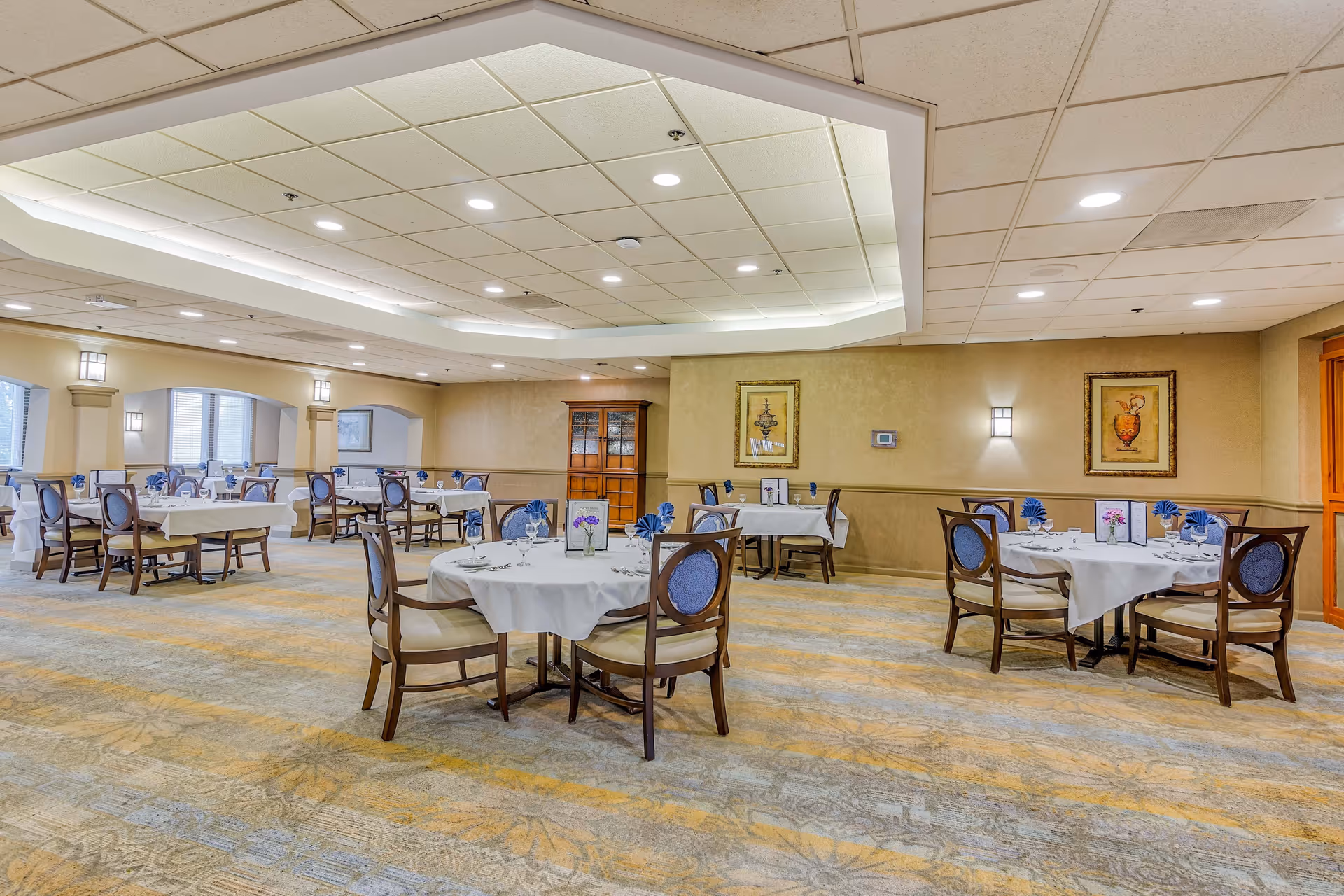 A spacious dining room with round and rectangular tables covered with white tablecloths, each set with glassware, napkins, and small floral centerpieces. The room has a patterned carpet, soft yellow walls with framed artwork, and recessed lighting in the ceiling. Wooden chairs with blue upholstered backs surround the tables.