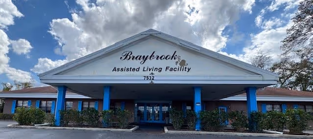 Front exterior view of Braybrook Assisted Living Facility showing a large building with a peaked roof, blue pillars, and bushes in front under a partly cloudy sky.
