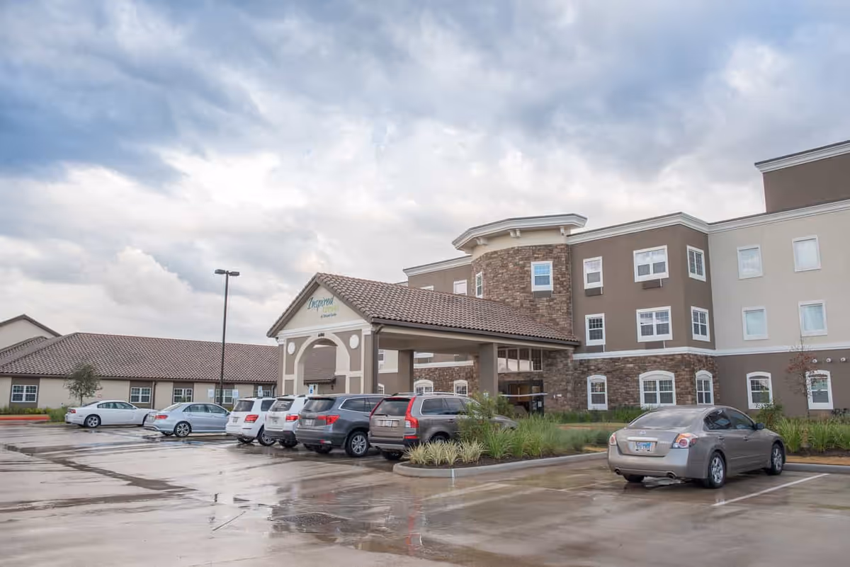 Exterior front entrance of a multi-story senior living facility with a covered porte-cochere and cars parked in a wet parking lot under a cloudy sky.